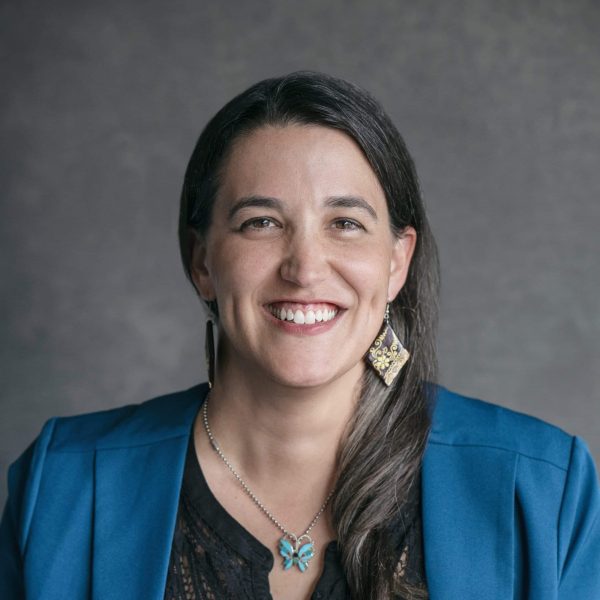 A woman with long dark hair wearing a blue blazer, black top, dangling earrings, and a butterfly necklace smiles in front of a gray background.