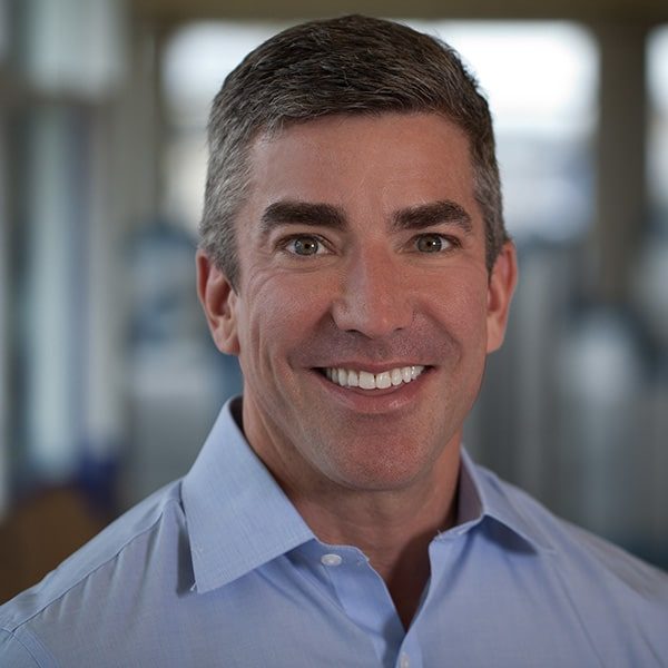 A Whitman Walker staff physician with short gray hair smiles at the camera, wearing a light blue collared shirt, with a blurred indoor background.