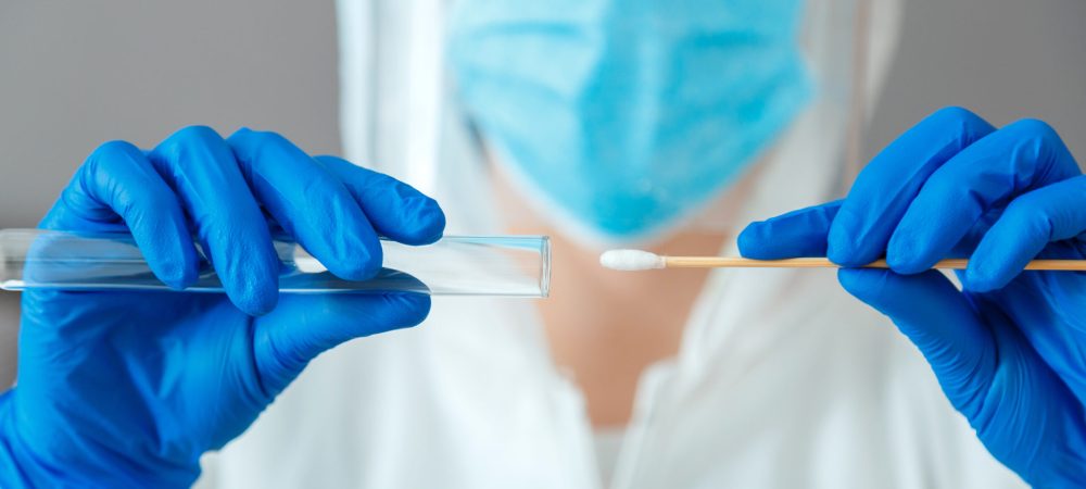 Person wearing a mask and gloves holds a test tube in one hand and a cotton swab in the other, preparing for COVID-19 testing and sample collection.