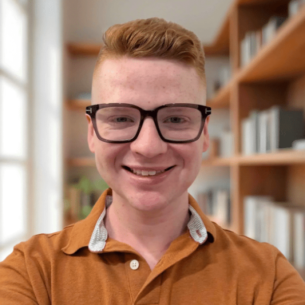 A person wearing glasses and a brown collared shirt smiles at the camera in a room with bookshelves and natural light.