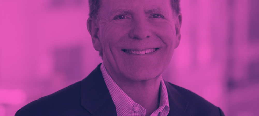 Man in a suit jacket and checkered shirt smiling at the camera, with an out-of-focus building in the background under a pink color filter.