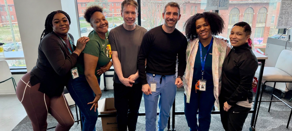 Six people stand closely together indoors, smiling at the camera in front of large windows with a view of buildings outside.