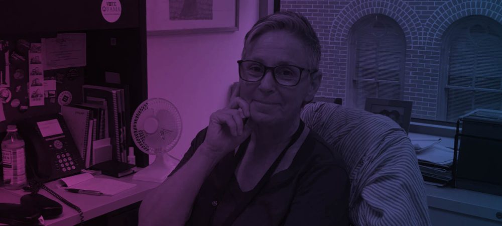 A person with short hair and glasses sits at an office desk, resting their hand on their chin, with shelves, a fan, and a window in the background.