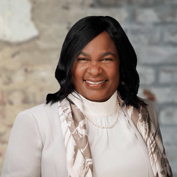 A woman in a light-colored blazer and patterned scarf smiles in front of a blurred brick wall background, embodying Whitman Walker leadership with confidence and warmth.