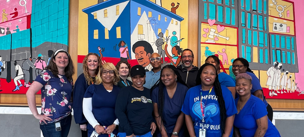 A group of twelve people pose and smile in front of two colorful, illustrated murals depicting community scenes and buildings, highlighting the importance of women’s health in the community.