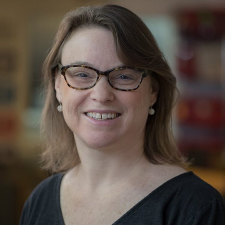A woman with shoulder-length brown hair, glasses, and pearl earrings smiles at the camera. She is wearing a dark top and standing indoors with a blurred background, reflecting her role as Whitman Walker Executive Director.