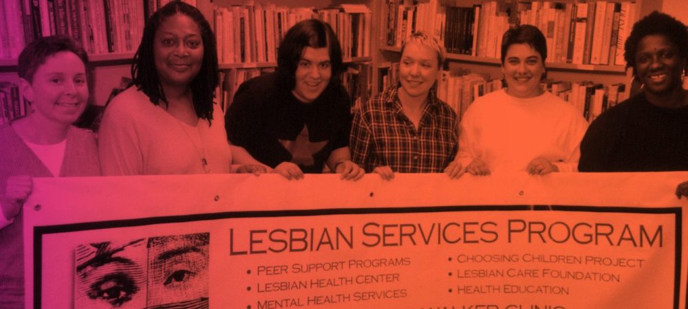 Six people stand together in front of bookshelves, holding a banner that reads “Lesbian Services Program” with a list of support and health services.
