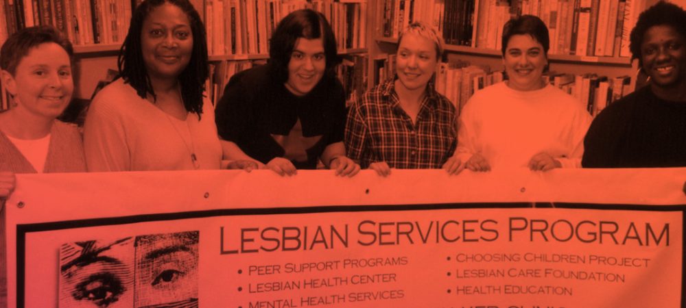 Six people stand behind a banner for the Lesbian Services Program at Whitman-Walker Clinic, advertising peer support, health services, and a contact phone number. Bookshelves are in the background.