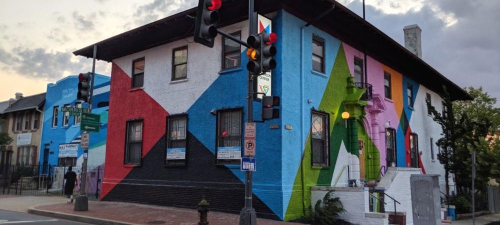 A two-story corner building with a vibrant geometric mural in red, black, blue, green, and white, next to a traffic light and street signs.