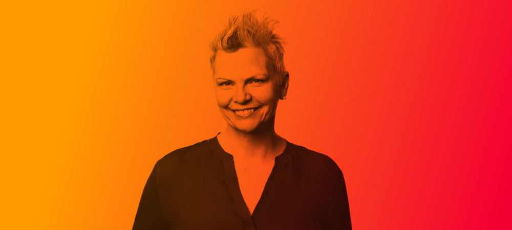 Person with short, spiked hair and a dark shirt smiling at the camera, set against a vibrant orange and red gradient background.