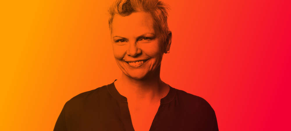 Smiling person with short, spiked hair wearing a dark shirt, standing in front of a gradient orange to red background.