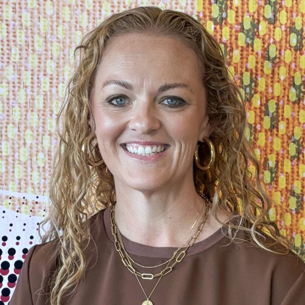 A woman with curly blonde hair, wearing gold hoop earrings, layered necklaces, and a brown top, smiling in front of a colorful patterned background.