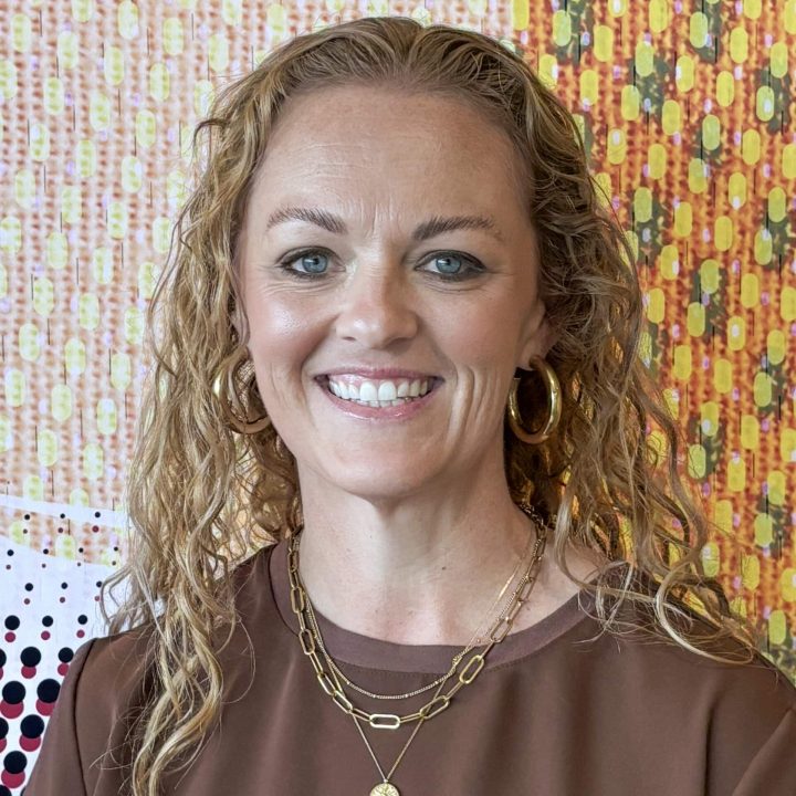 A woman with curly blonde hair, wearing gold hoop earrings, layered necklaces, and a brown top, smiling in front of a colorful patterned background.