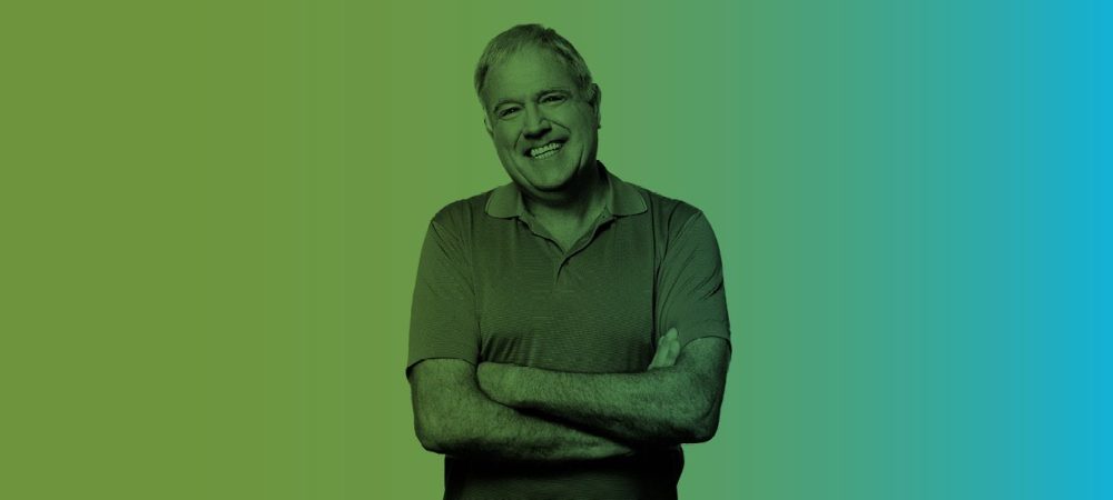 A middle-aged man in a short-sleeve polo shirt stands with arms crossed, smiling, against a green to blue gradient background.