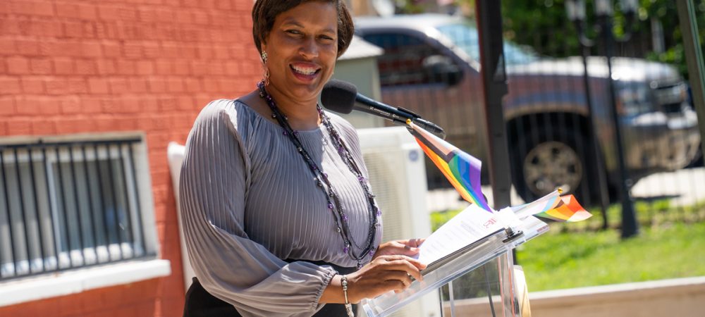 A woman stands at a podium with small rainbow flags, smiling and speaking into a microphone at an outdoor event.