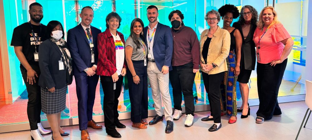A group of eleven people stand together indoors, smiling at the camera in front of a glass wall with colorful lighting during the NIH NIAID visit to Max Robinson Center.