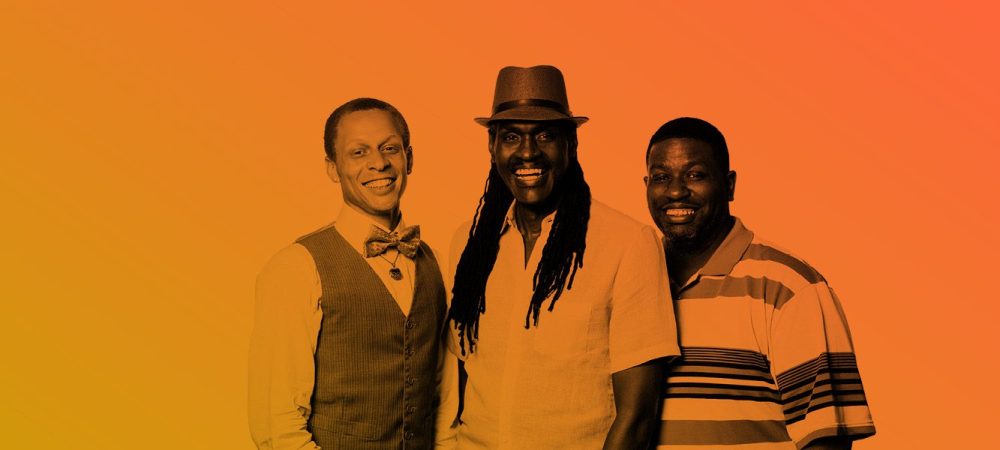 Three men stand side by side, smiling at the camera; one wears a vest and bow tie, another a hat and casual shirt, and the third a striped polo shirt. The background is orange gradient.