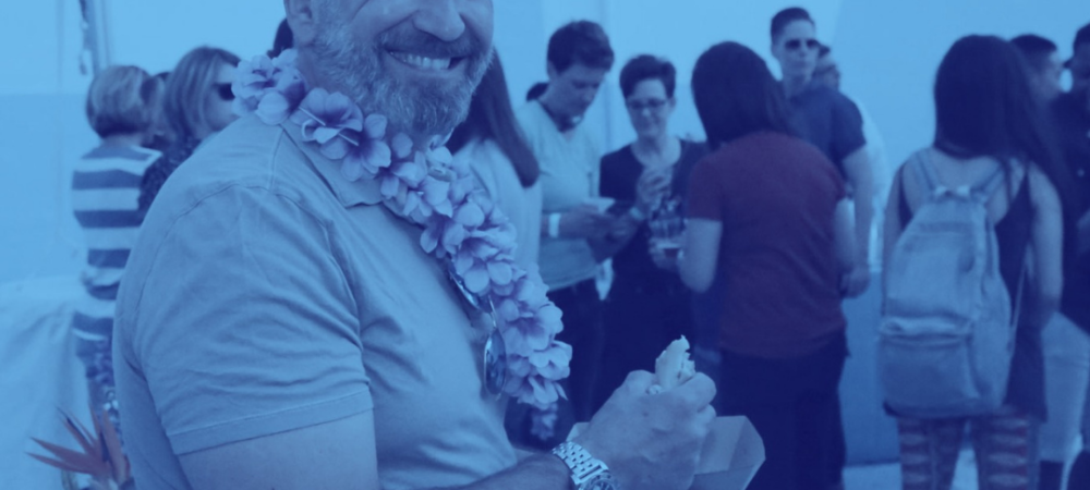 A man wearing a floral lei smiles at the camera while holding food at an outdoor gathering with people socializing in the background.
