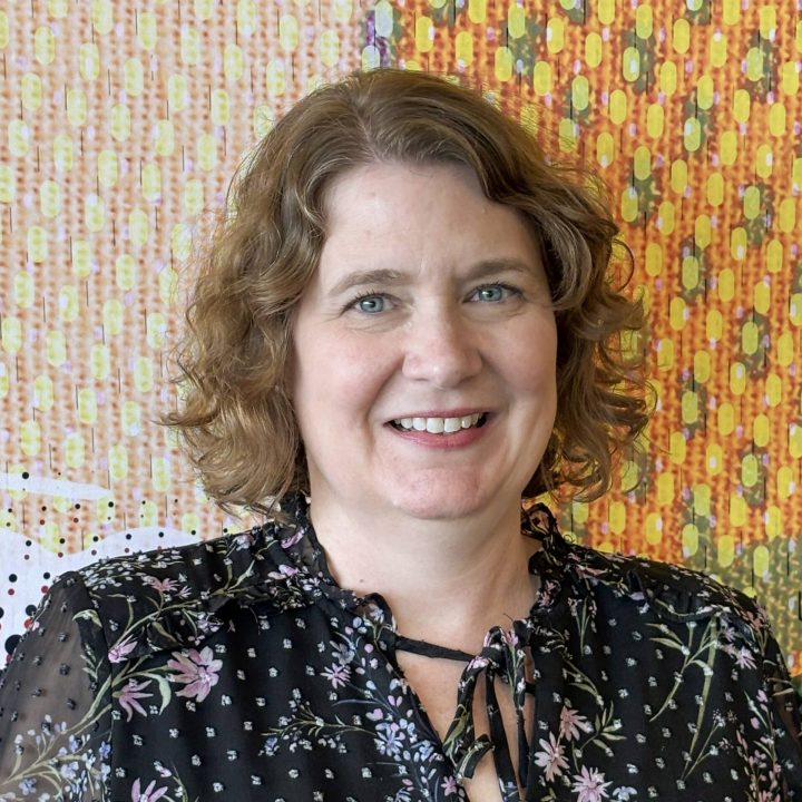 A woman with curly light brown hair, wearing a black floral blouse, smiles in front of a colorful, patterned background.