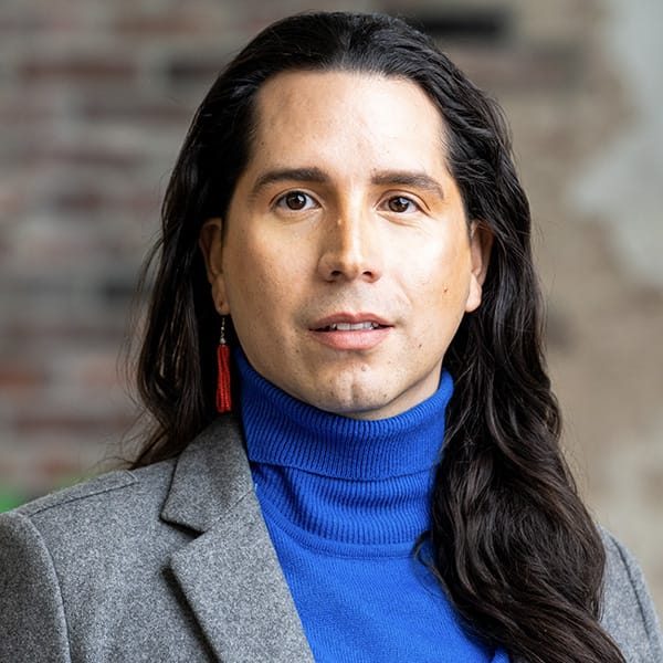 Person with long dark hair wearing a gray blazer, blue turtleneck, and red earring, standing in front of a blurred brick and cream background.