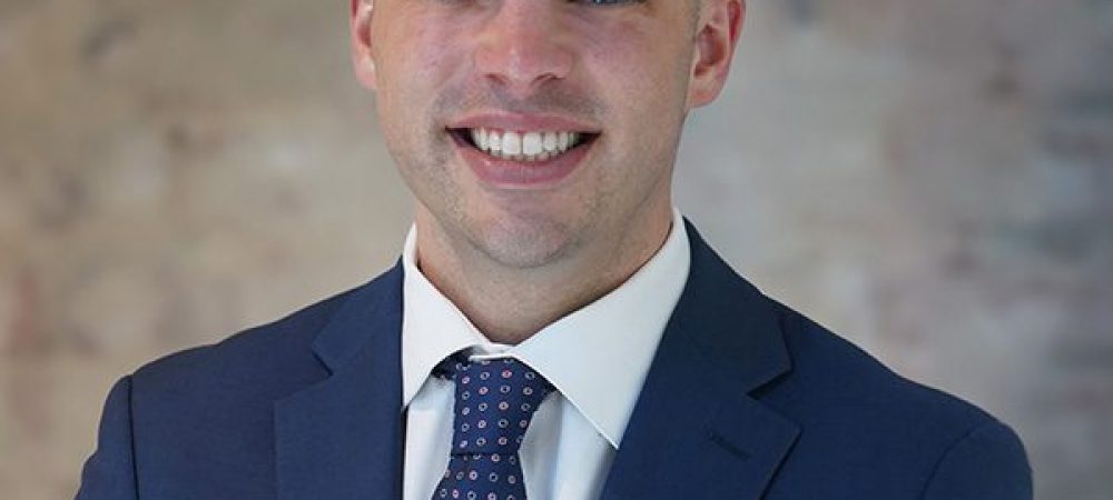 A CEO in a blue suit, white shirt, and patterned tie stands smiling in front of a textured, light-colored wall.