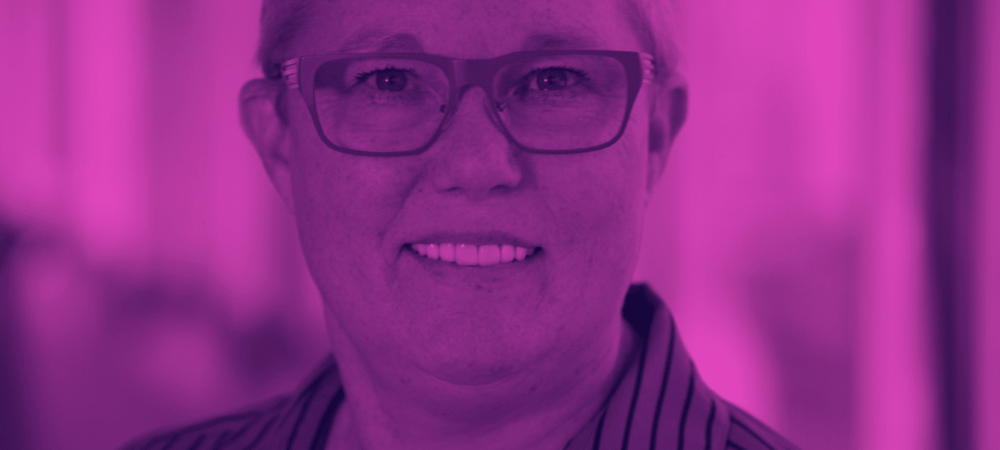 A person with short hair and glasses, wearing a striped shirt and a heart-shaped necklace, smiles at the camera. The image has a purple color overlay.
