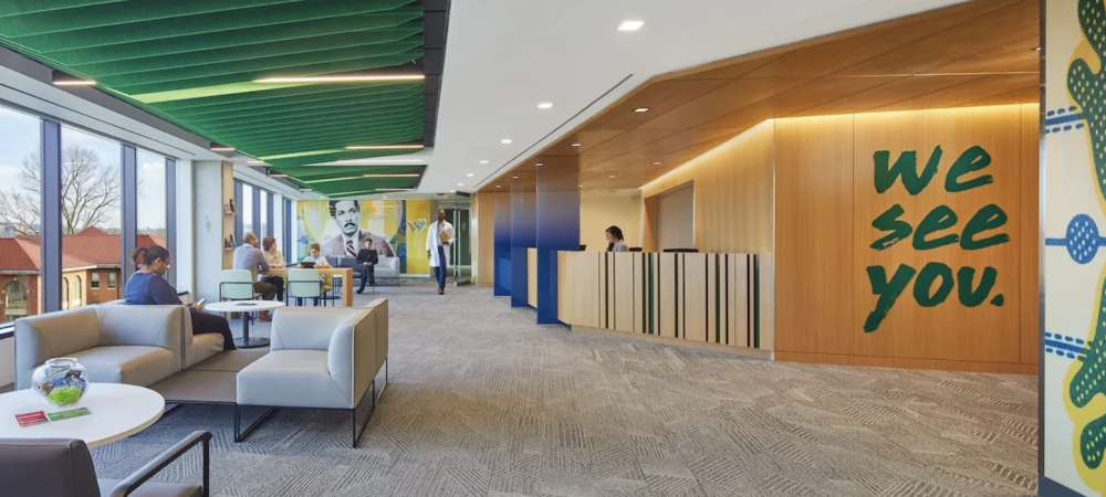 Modern office lobby with seating area, green ceiling panels, wall art reading "we see you," reception desk, and large windows filled with natural light—a welcoming Whitman Walker Congress Heights celebration of community.
