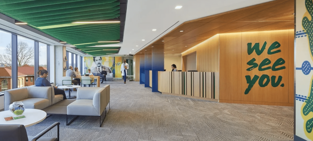 Modern office lobby with seating area, green ceiling panels, wall art reading "we see you," reception desk, and large windows filled with natural light—a welcoming Whitman Walker Congress Heights celebration of community.