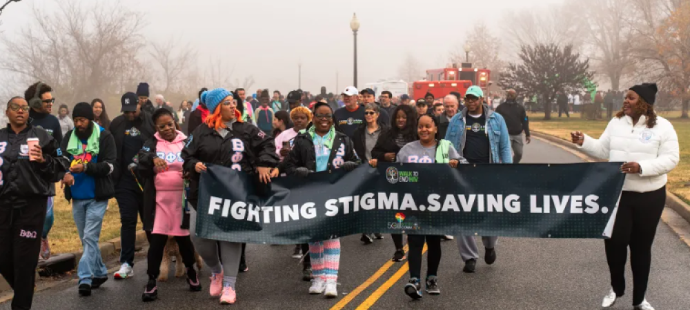 A diverse group of people march outdoors on a foggy day at the Walk to End HIV 2024 in Anacostia Park, holding a banner that reads, "Fighting stigma. Saving lives.