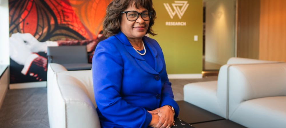 A woman in a blue blazer sits on a light-colored sofa in a modern office with "RESEARCH" and a geometric logo on a green wall in the background.