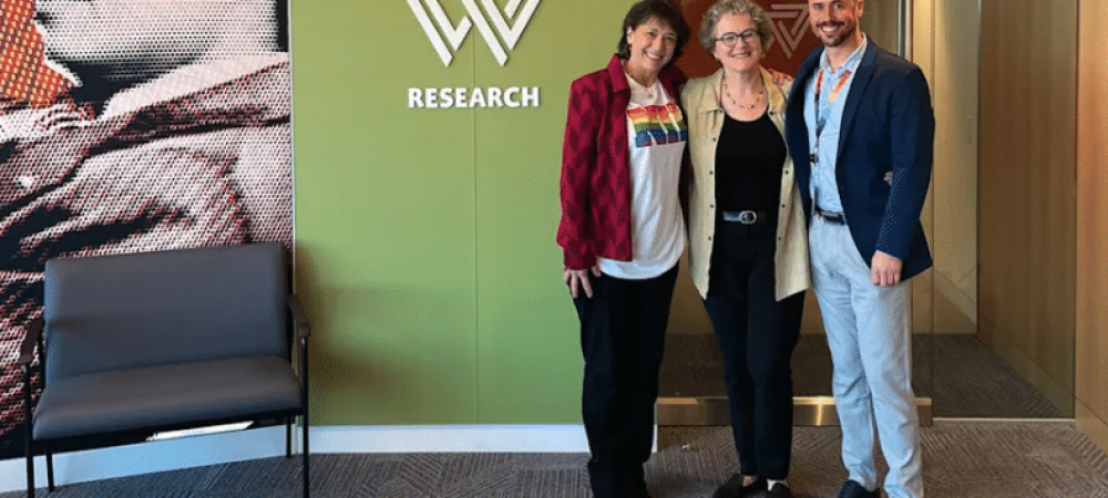 Three people stand together, smiling, in front of a wall with a "RESEARCH" sign and logo; their gathering highlights concerns about HIV research funding cuts in the USA. A chair and partial mural are visible on the left.