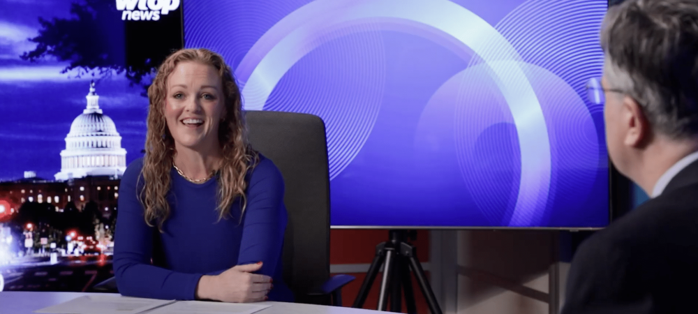 A woman in a blue dress sits at a news desk smiling, with a man seated across from her and the U.S. Capitol displayed on a screen behind them.