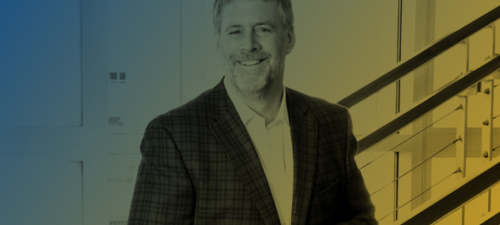 A man in a checked blazer stands smiling by a modern staircase, with a gradient blue and yellow filter over the image.