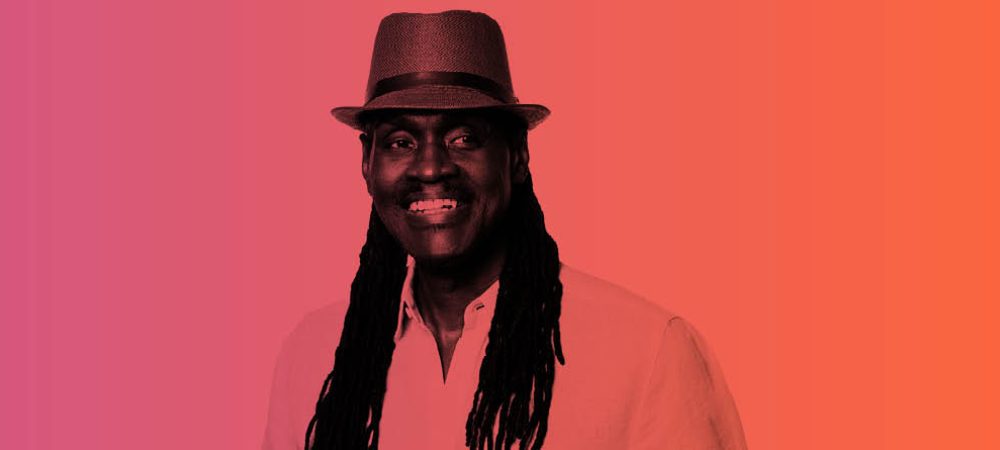 A smiling person with long dreadlocks and a hat stands against a gradient background of pink, purple, and orange hues.