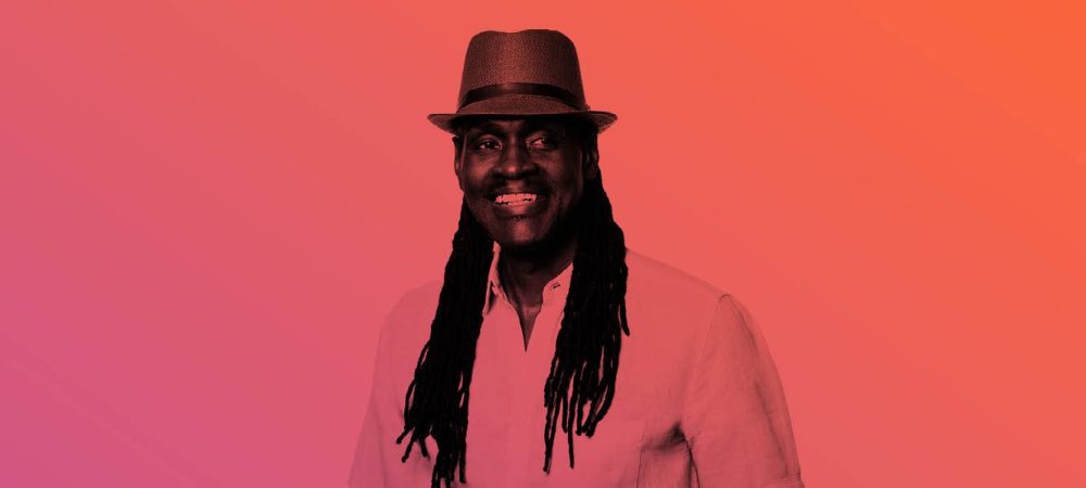 A man wearing a light shirt and a fedora hat, smiling, with long dreadlocks, standing against a pink and orange gradient background.