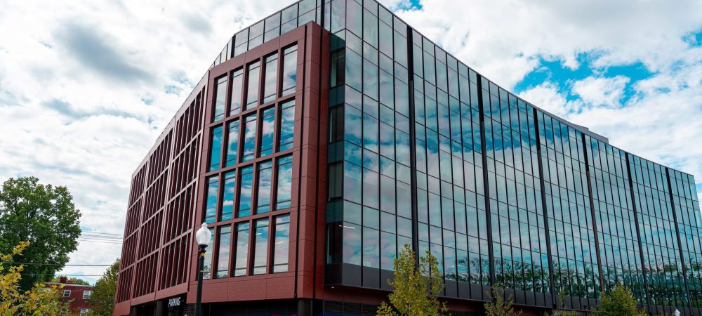 A modern glass office building with a red-brown frame, seen from the street corner, with cars parked and people walking on the sidewalk.
