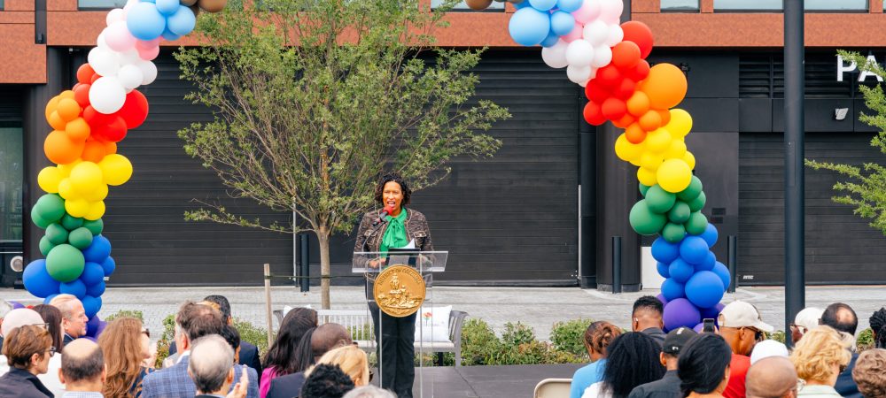 A woman speaks at a podium under a colorful balloon arch in front of an audience at the Max Robinson Center 1st anniversary outdoor event.