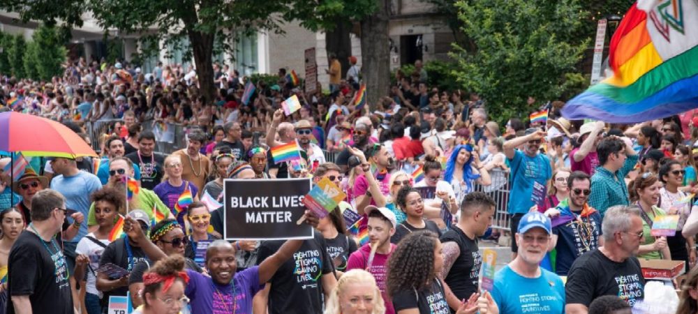 A large crowd participates in a Pride parade, with people holding rainbow flags and a sign reading "Black Lives Matter.