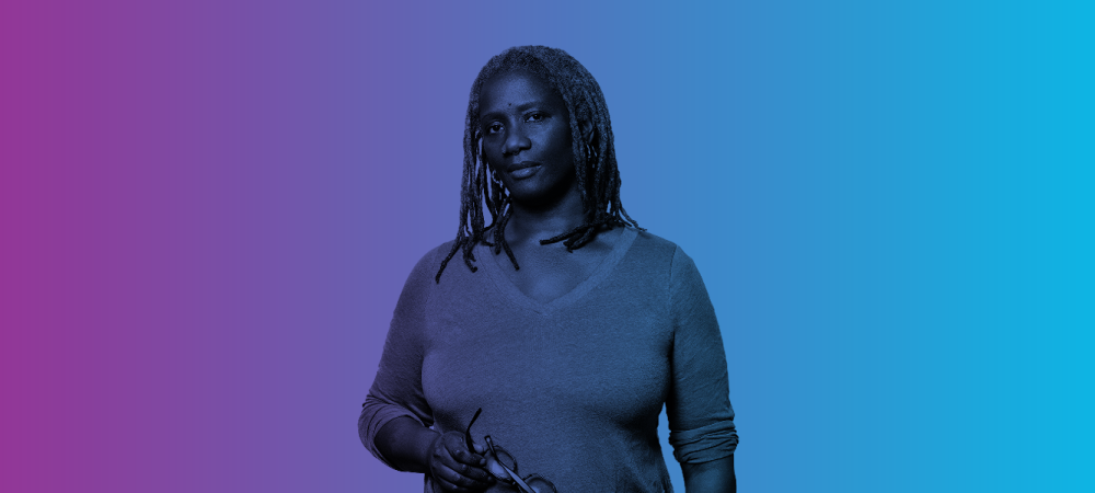 A person with long, gray dreadlocks wearing a sweater holds eyeglasses and stands against a purple-to-blue gradient background.