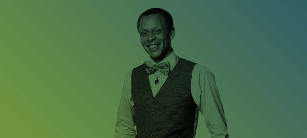 A man wearing a bow tie, vest, and collared shirt stands smiling against a gradient green to blue background.