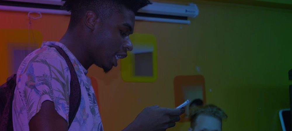 A person with a backpack reads from a phone while another person with a beard looks on in a room with colorful square decorations on the wall.
