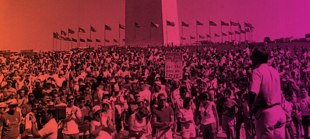A large crowd gathers in front of the Washington Monument, with a person addressing them and American flags in the background.