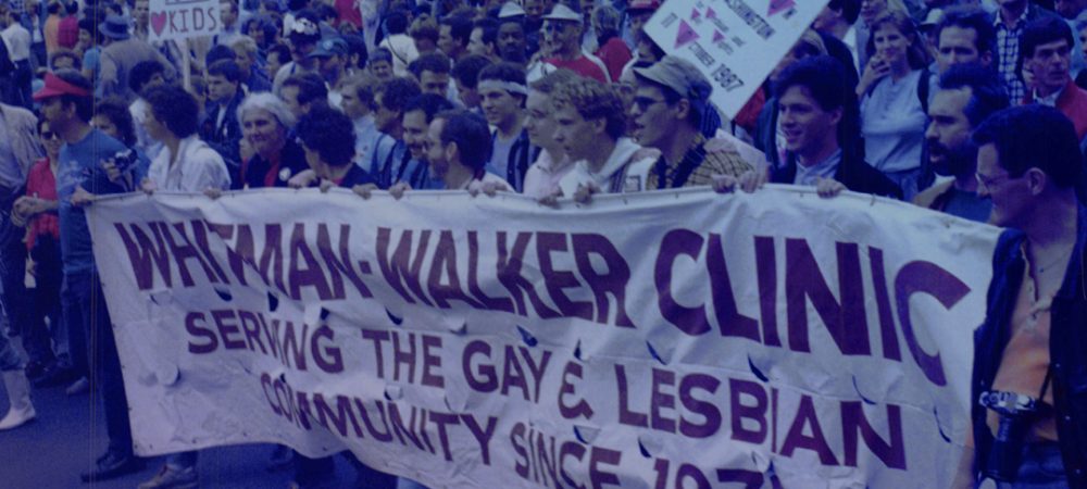 A large group of people march holding a banner that reads “Whitman-Walker Clinic Serving the Gay & Lesbian Community Since 1973.” Signs and a crowd are visible in the background.