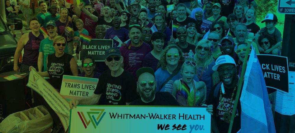 A diverse group of people at a pride event hold signs reading "Black Lives Matter" and "Trans Lives Matter" near a Whitman-Walker Health banner.