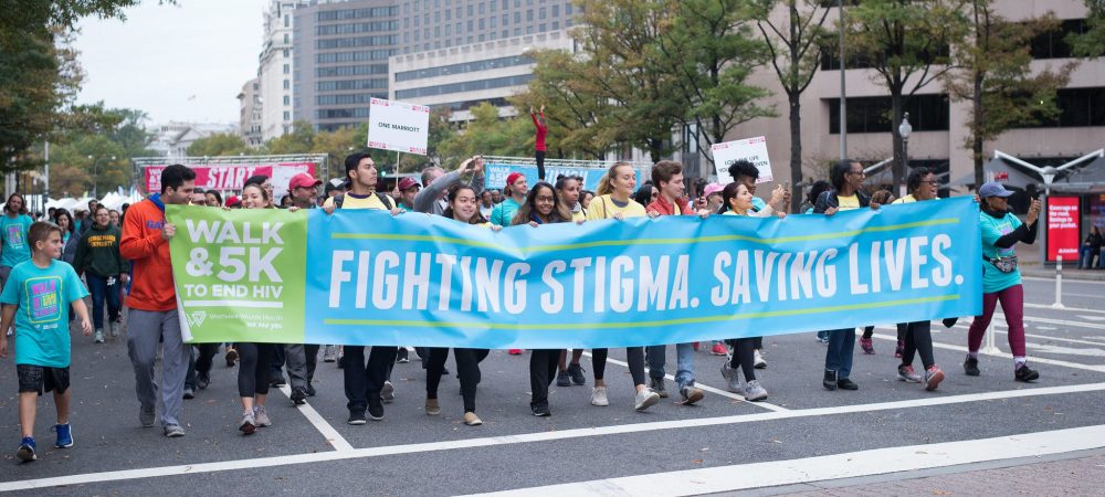 A group of people walk in a city street holding banners that read "Fighting Stigma. Saving Lives." and "Walk & 5K to End HIV.