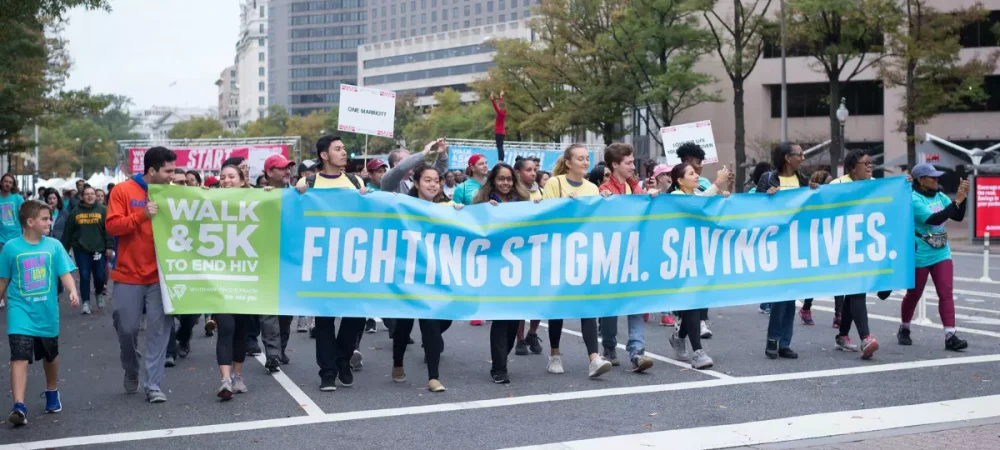 A group of people walk in a city street holding a large banner that reads "Fighting Stigma. Saving Lives." at a Walk & 5K to End HIV event.