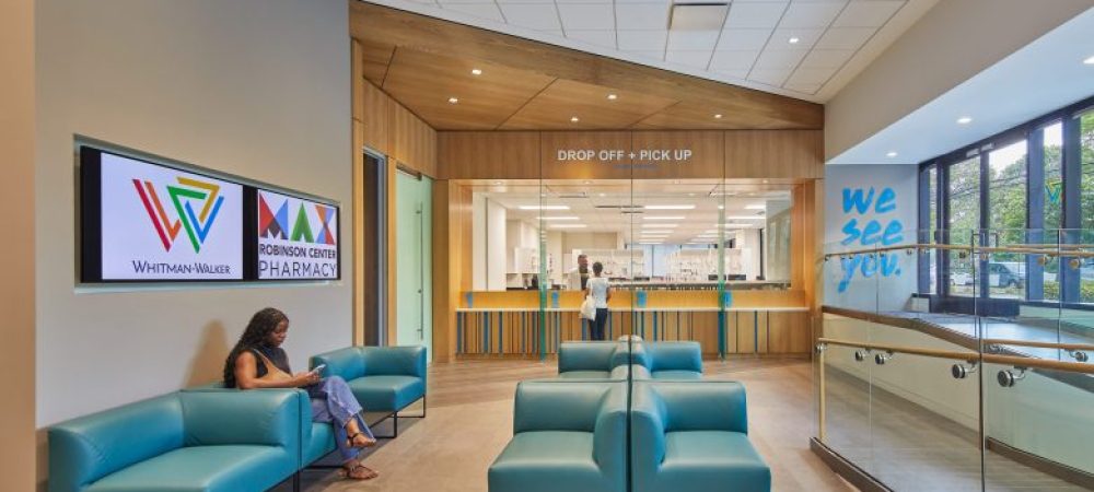 A woman sits on a teal chair in a modern pharmacy waiting area with signage, large windows, and a counter labeled "Drop Off + Pick Up.
