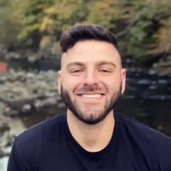 A man with short dark hair and a beard is smiling, standing outdoors near a rocky river with trees in the background.