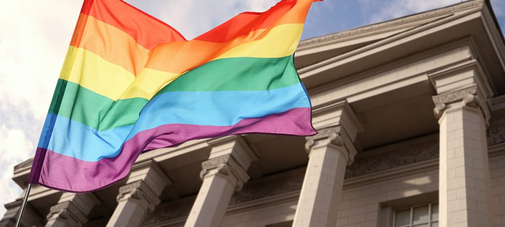 A rainbow pride flag waves in front of a classical-style building with large columns under a partly cloudy sky.