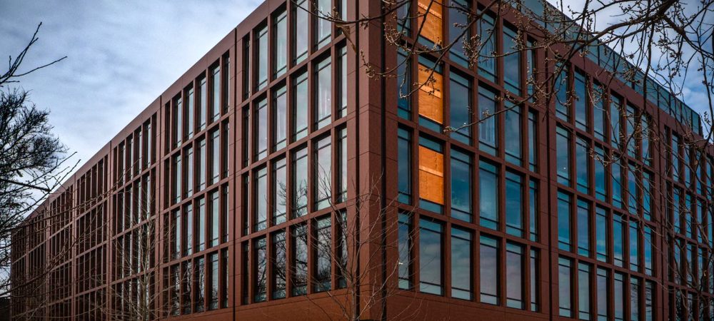 A modern, multi-story office building with large glass windows and a reddish-brown exterior, surrounded by leafless trees under a cloudy sky.